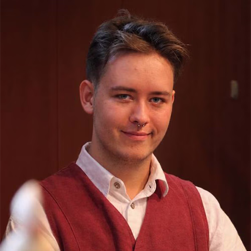 Young person with short, styled hair and a septum piercing wearing a white shirt and a red vest, smiling slightly against a dark background.