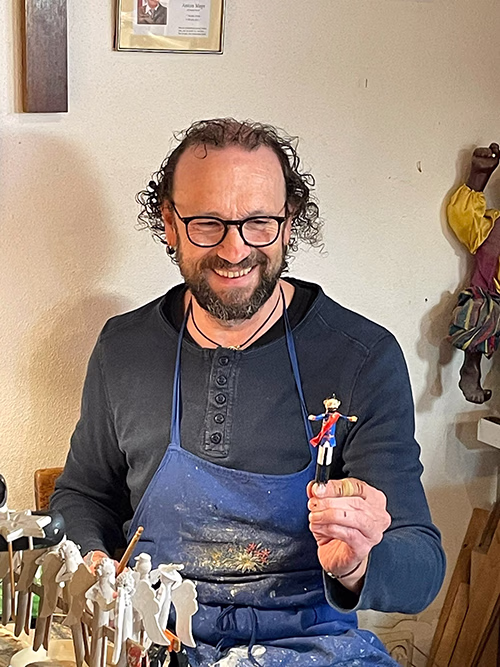 Smiling man with glasses and curly hair wearing a dark shirt and a blue apron, holding a small figurine of a soldier wearing a red cape and blue uniform, sitting in a workspace with several similar figurines on sticks in front of him.