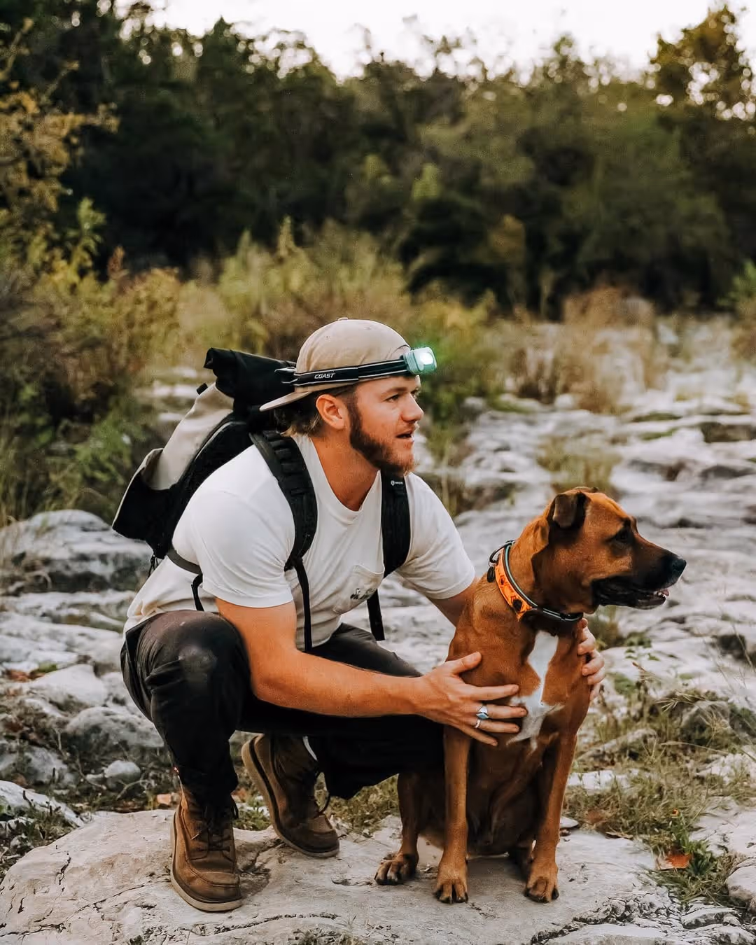 Man wearing a headlamp and backpack crouching on rocks, holding a brown dog with an orange collar, in an outdoor setting with trees.
