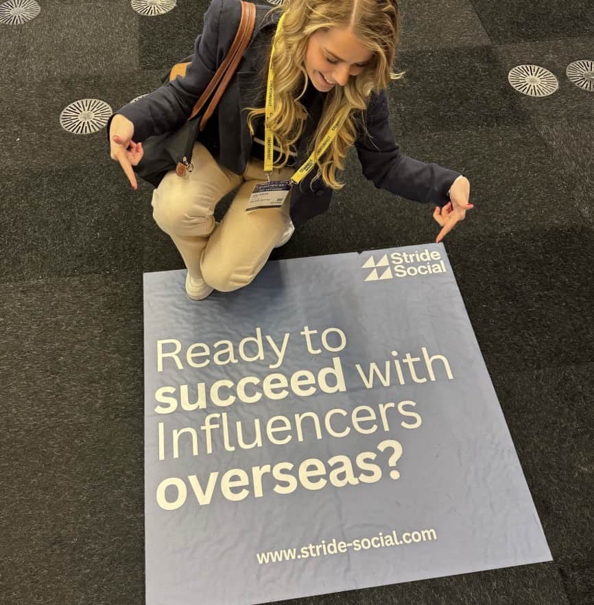 Woman crouching indoors pointing at a floor sign that reads 'Ready to succeed with Influencers overseas?' with Stride Social logo and website www.stride-social.com.
