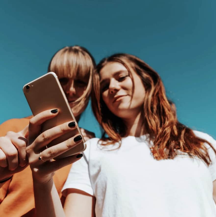 Two young women looking at a smartphone against a clear blue sky.