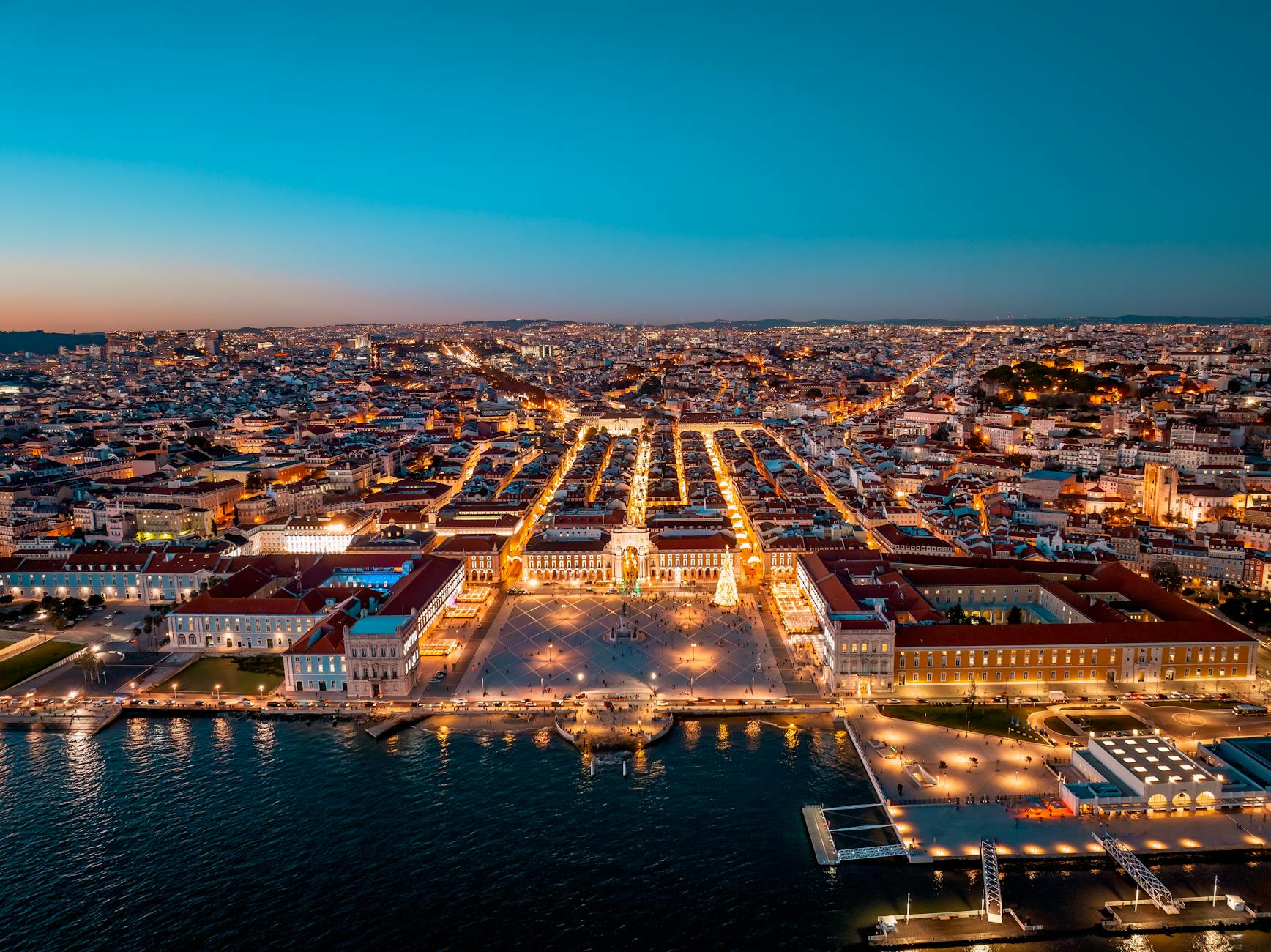 A stunning aerial view of Lisbon's illuminated cityscape and waterfront, captured at dusk.