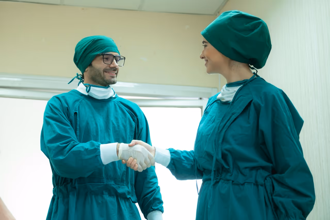 Two ENT medical professionals in green surgical scrubs and caps shaking hands and smiling.
