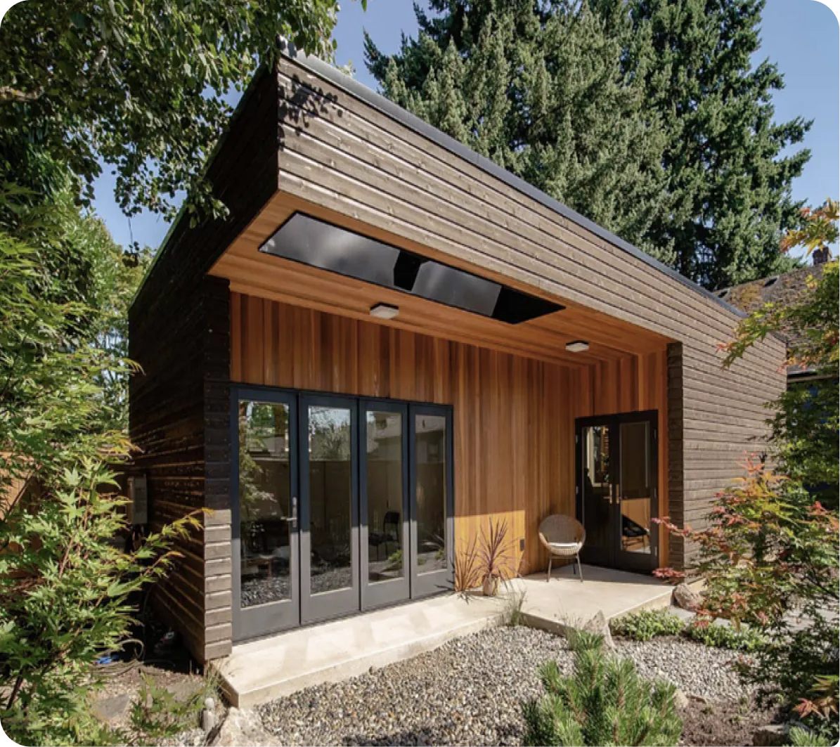 Modern wooden cabin with dark roof, glass doors, surrounded by tall evergreen trees and landscaping.
