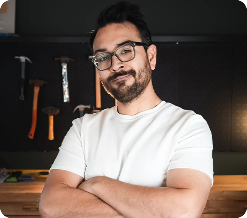 Man with glasses and beard wearing white shirt, arms crossed in workshop setting