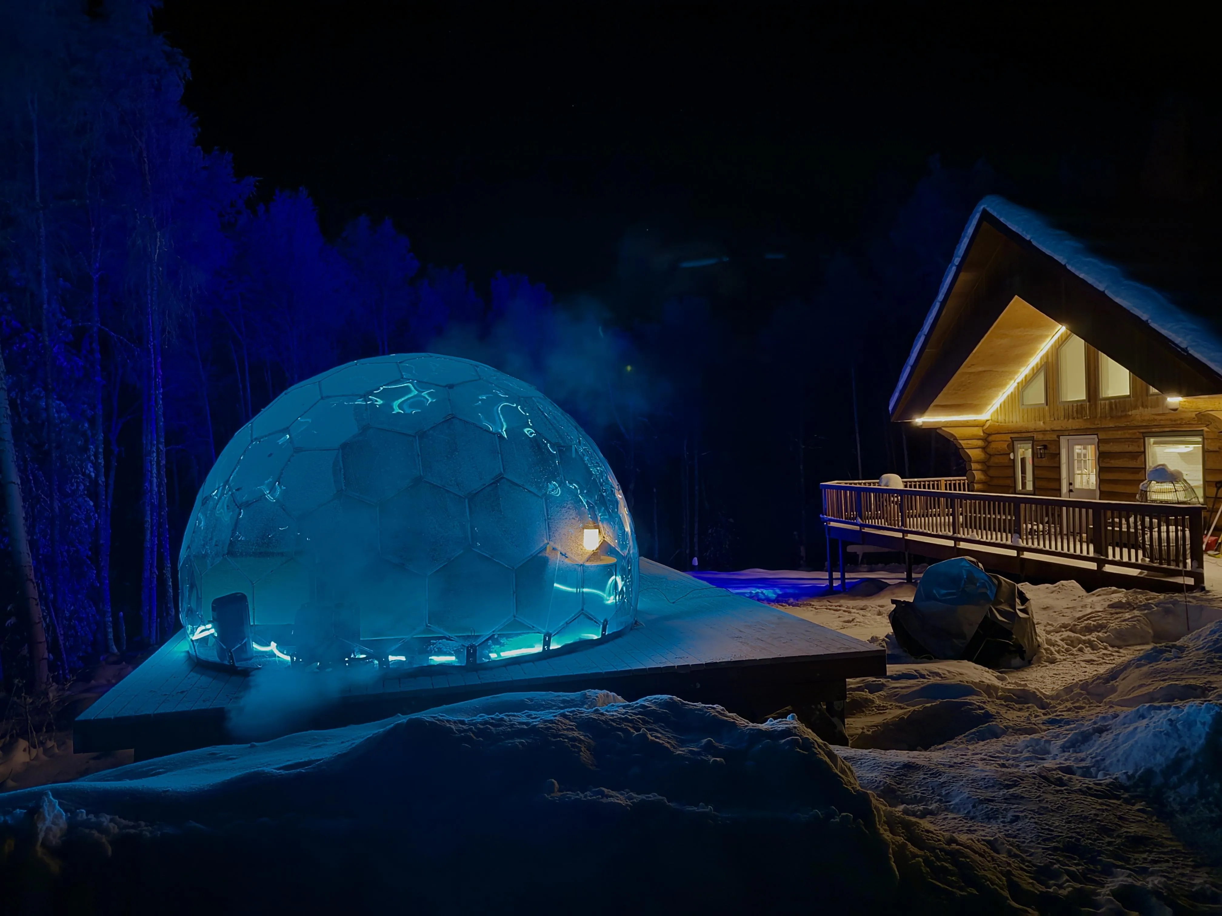 Illuminated geodesic dome and wooden cabin in snowy night landscape under blue lights
