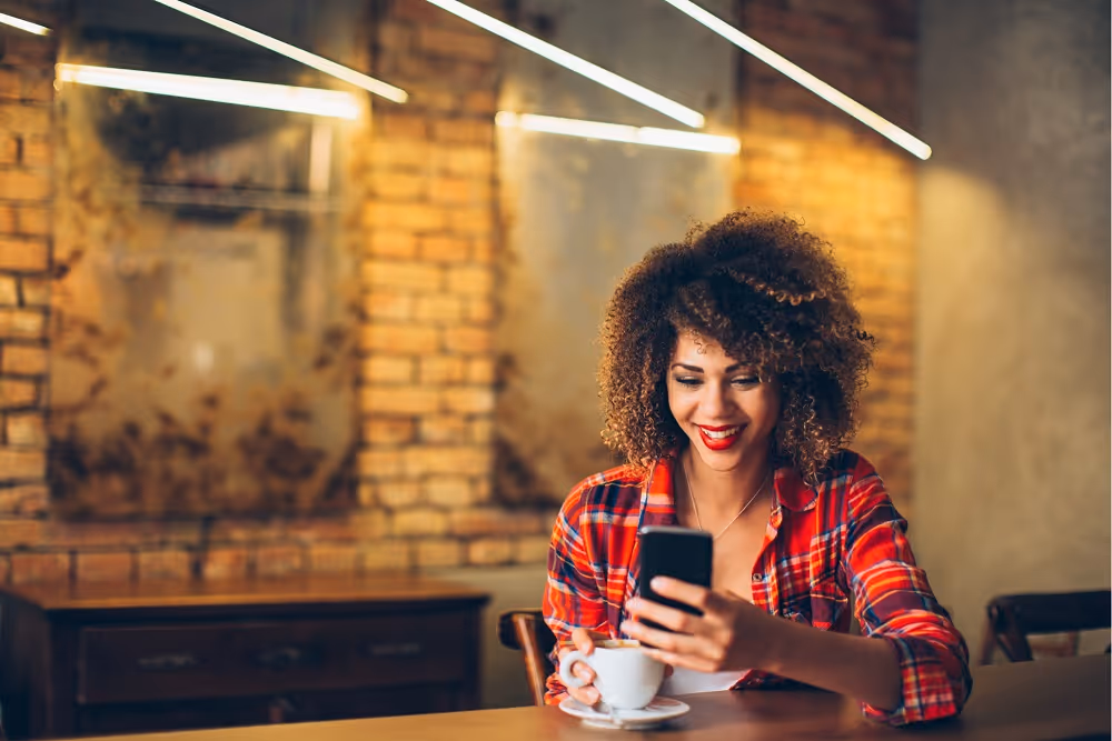 Smiling woman in red plaid shirt holding phone and cup, sitting in a cafe with brick walls.