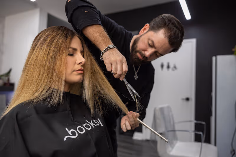 Hairdresser cutting a woman's long blonde hair in a salon wearing a black cape with 'booksy' logo.
