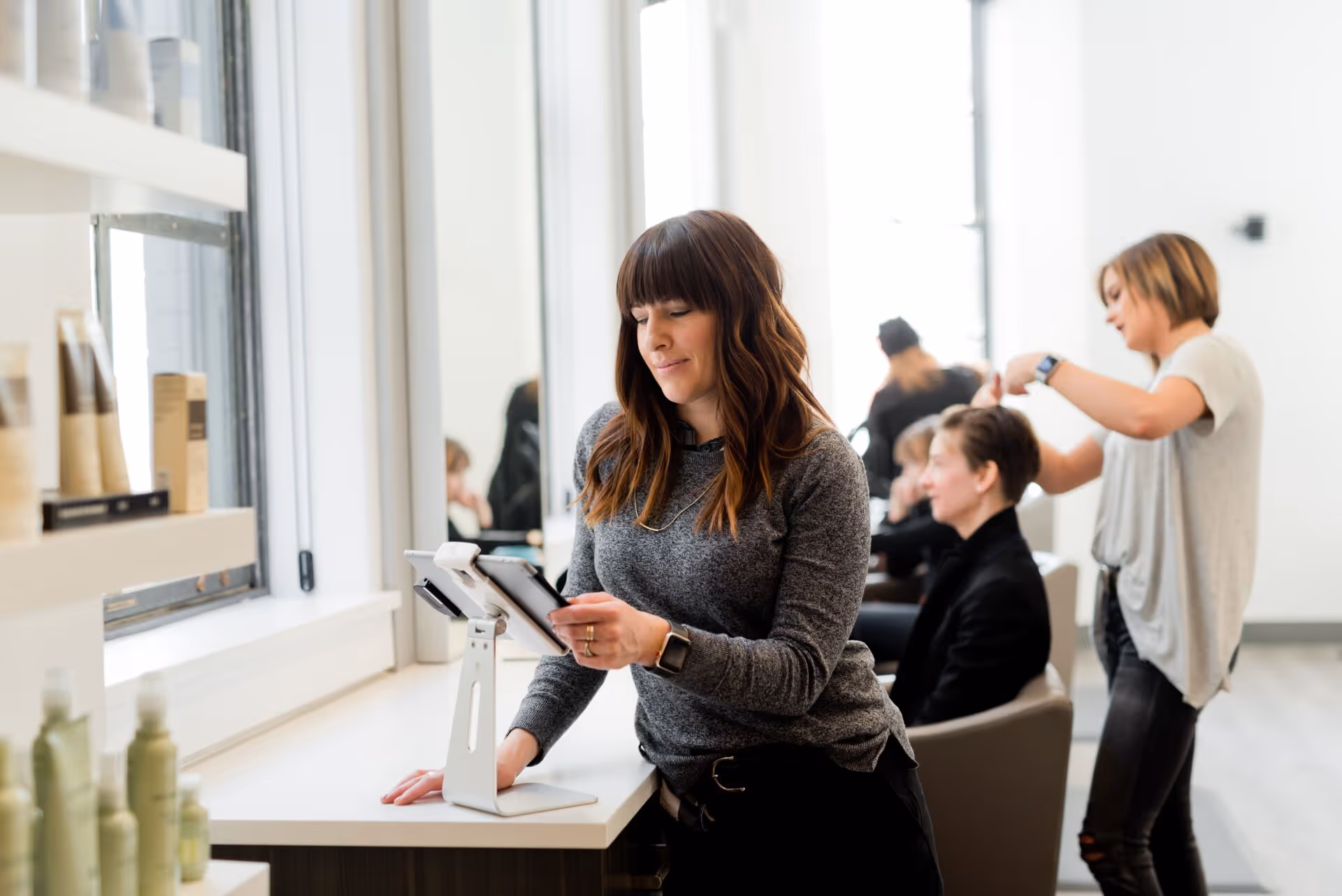 Woman standing at counter using tablet while hairstylist cuts a seated customer's hair in salon.