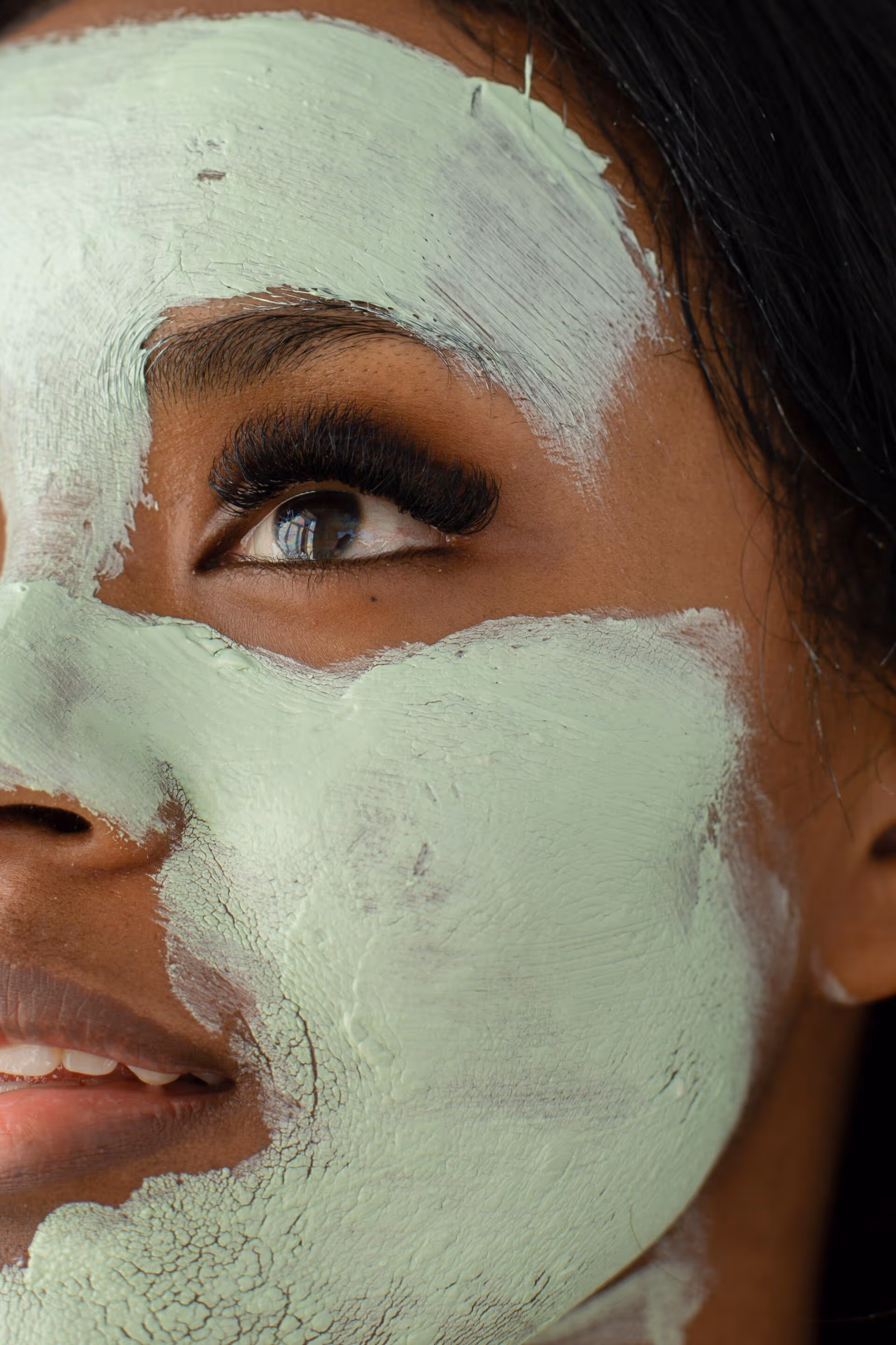 Close-up of a woman’s face with green facial mask applied and long eyelashes visible.