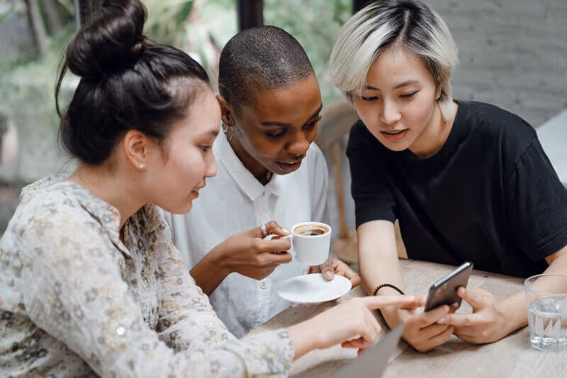 Three diverse women sitting at a table, looking at a smartphone and holding coffee cups.