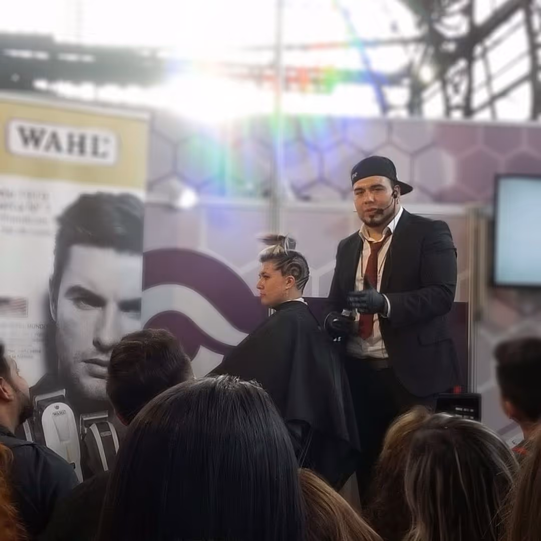 Barber in suit and cap giving a haircut demo to an audience under bright lights at a trade show booth.