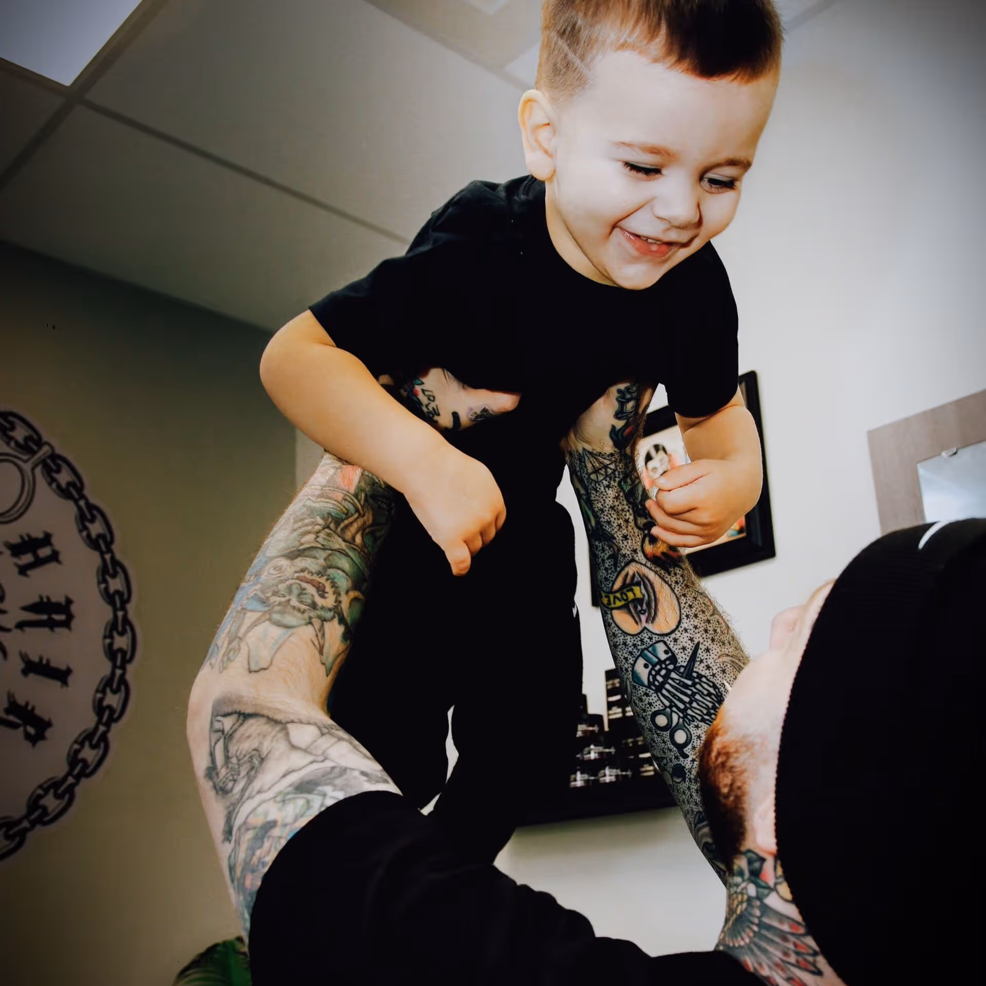 Tattooed adult lifting and smiling at a happy toddler wearing black clothes indoors.