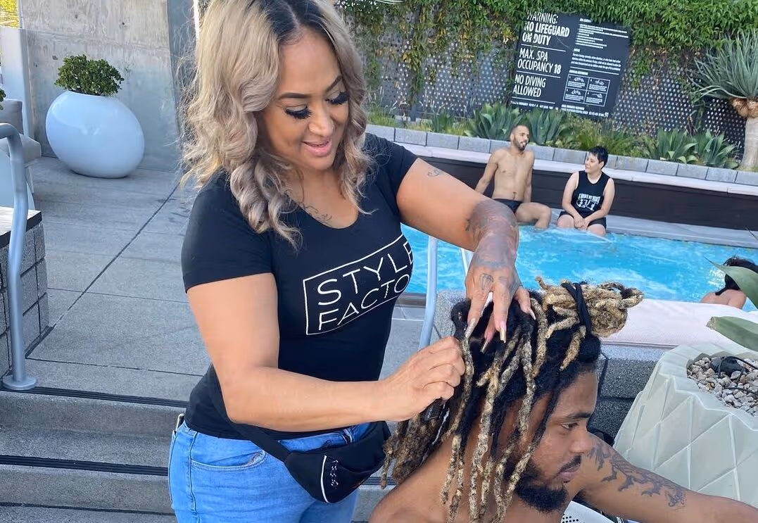 Woman styling man's long dreadlocks outdoors by a pool with people sitting in the background.