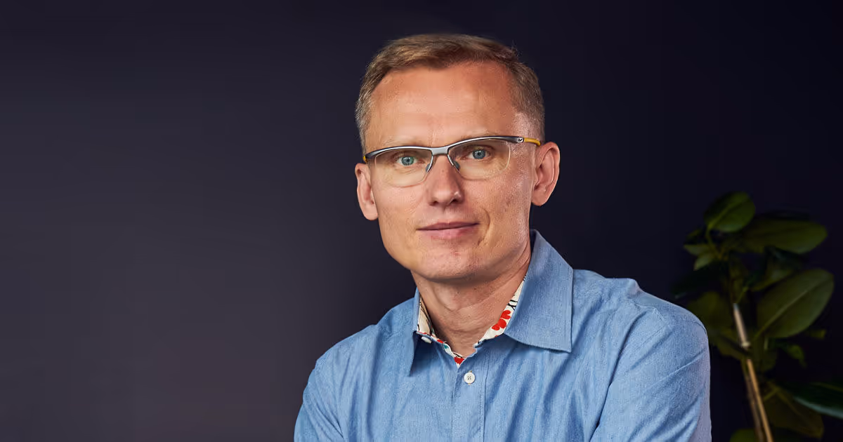 Man with short blond hair and glasses wearing a light blue shirt against a dark background.