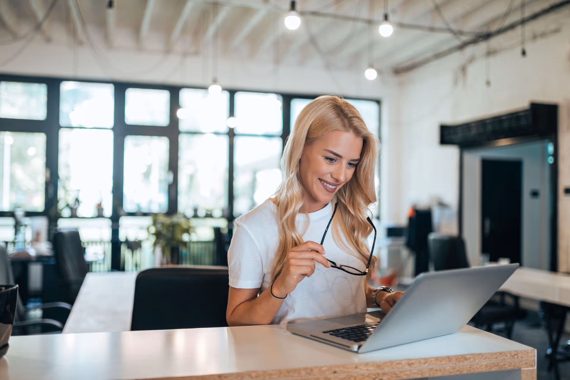 Smiling woman holding glasses while working on a laptop in a bright modern office.