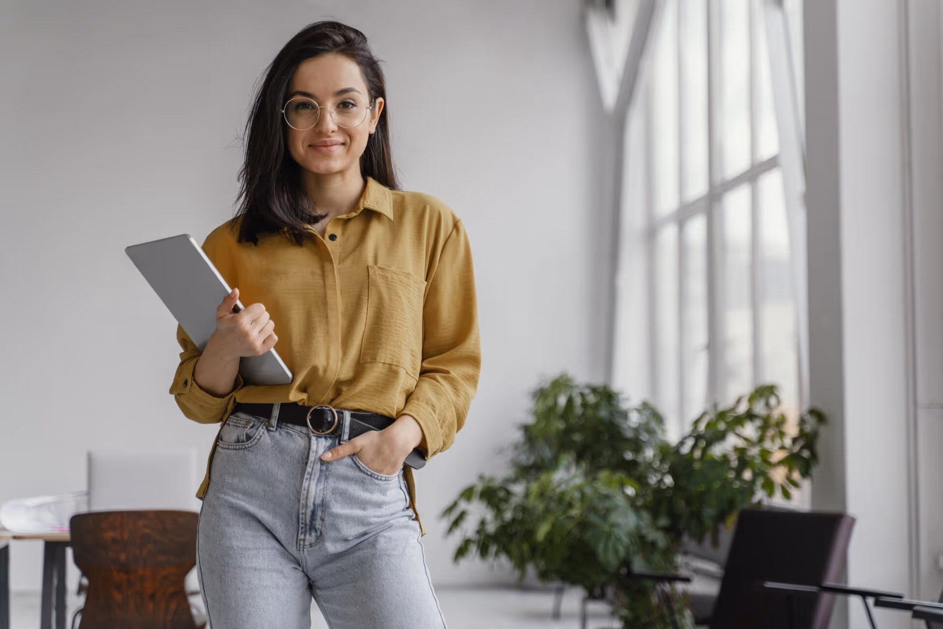 Young woman in glasses holding a tablet, standing in a bright office with plants and large windows.