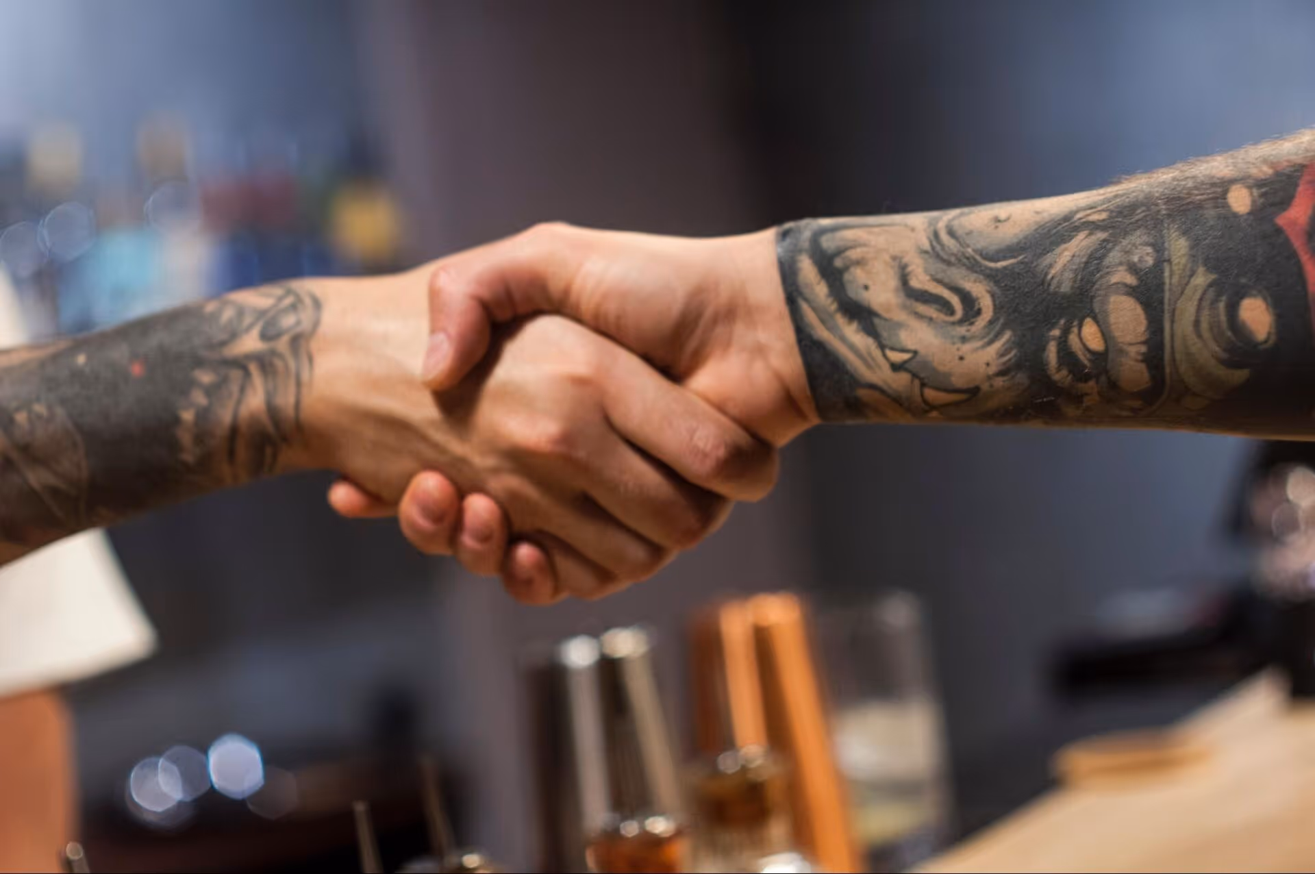 Two tattooed arms shaking hands in a close-up, blurred indoor background.