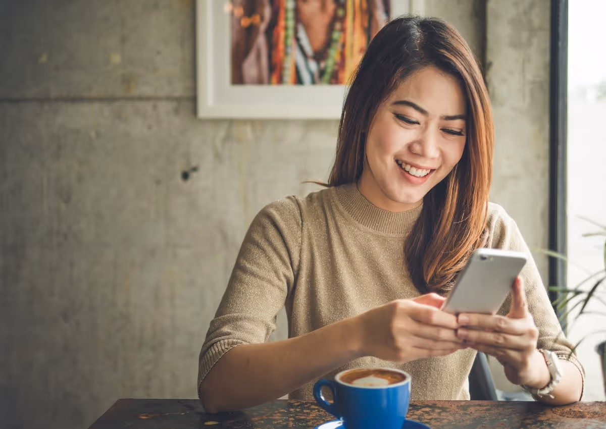 Smiling woman sitting at table, holding phone with a blue cup of coffee in front of her.