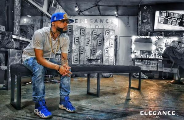 Man in blue cap, white shirt, and blue shoes sitting on bench inside barbershop with mirrors and chairs.