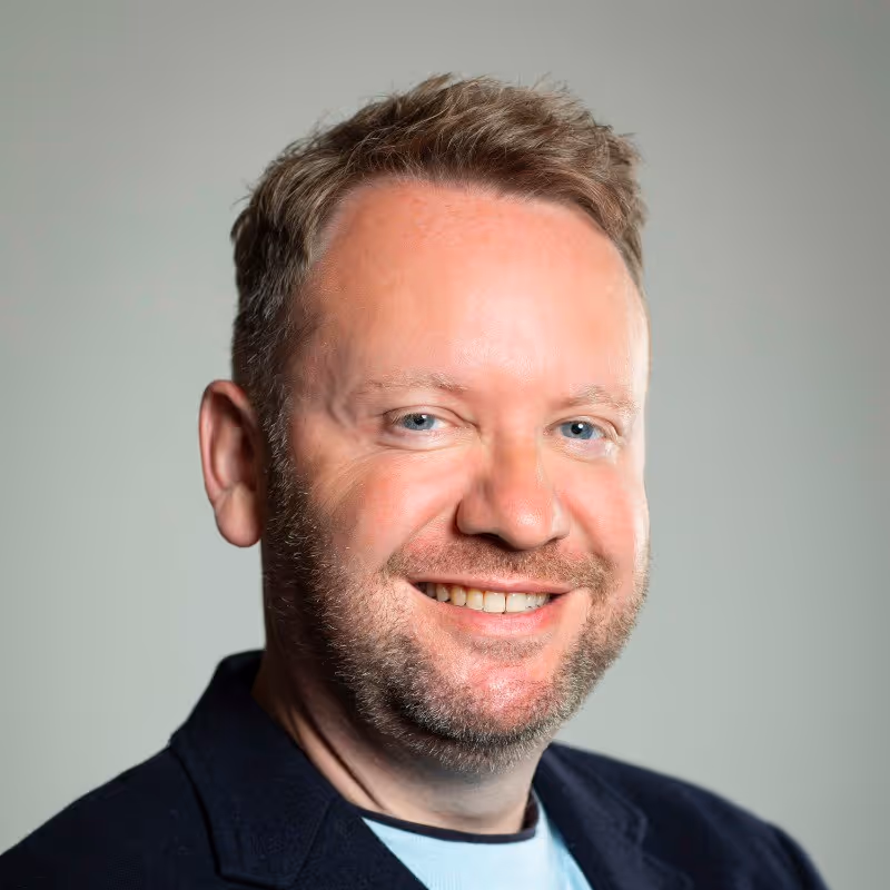Smiling man with short brown hair and beard wearing a light blue shirt and dark blazer.