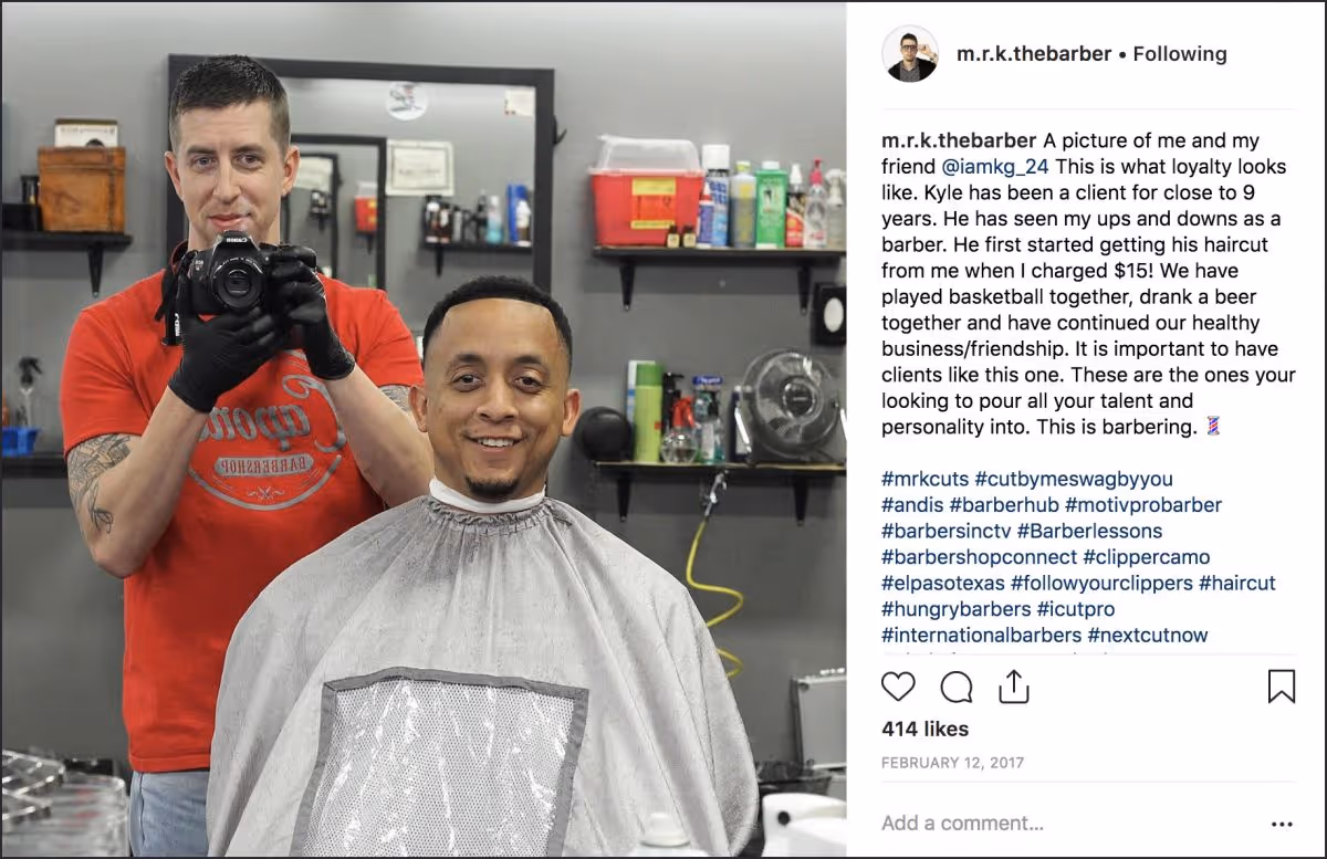 Barber in red shirt taking a photo in a mirror while client with fresh haircut smiles wearing a cape.