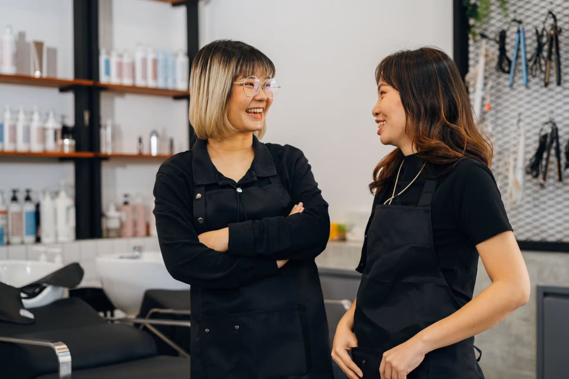 Two female hairstylists in black aprons smiling and chatting inside a modern salon.