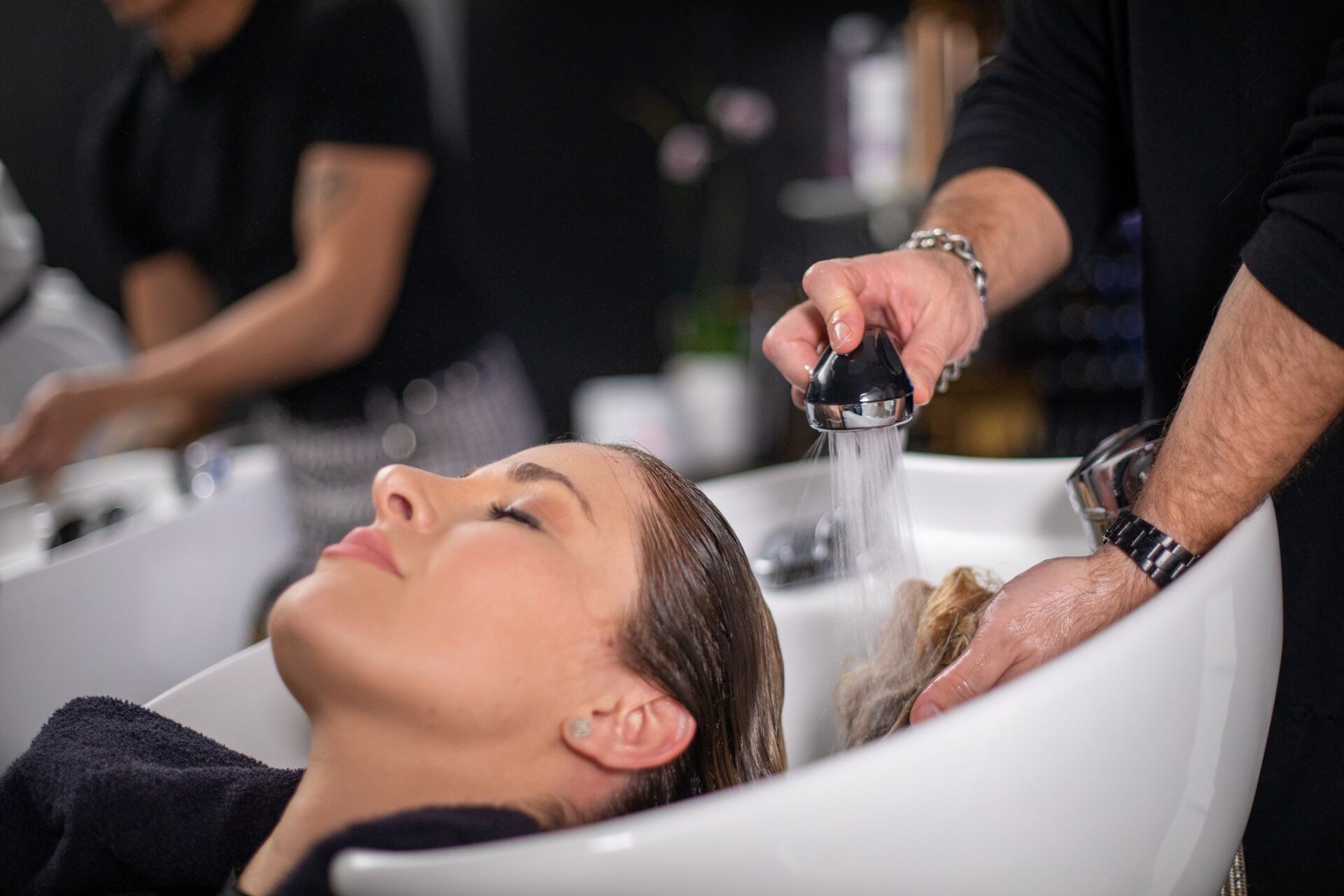 Woman with eyes closed having her hair washed in a salon sink with water flowing from a handheld sprayer.