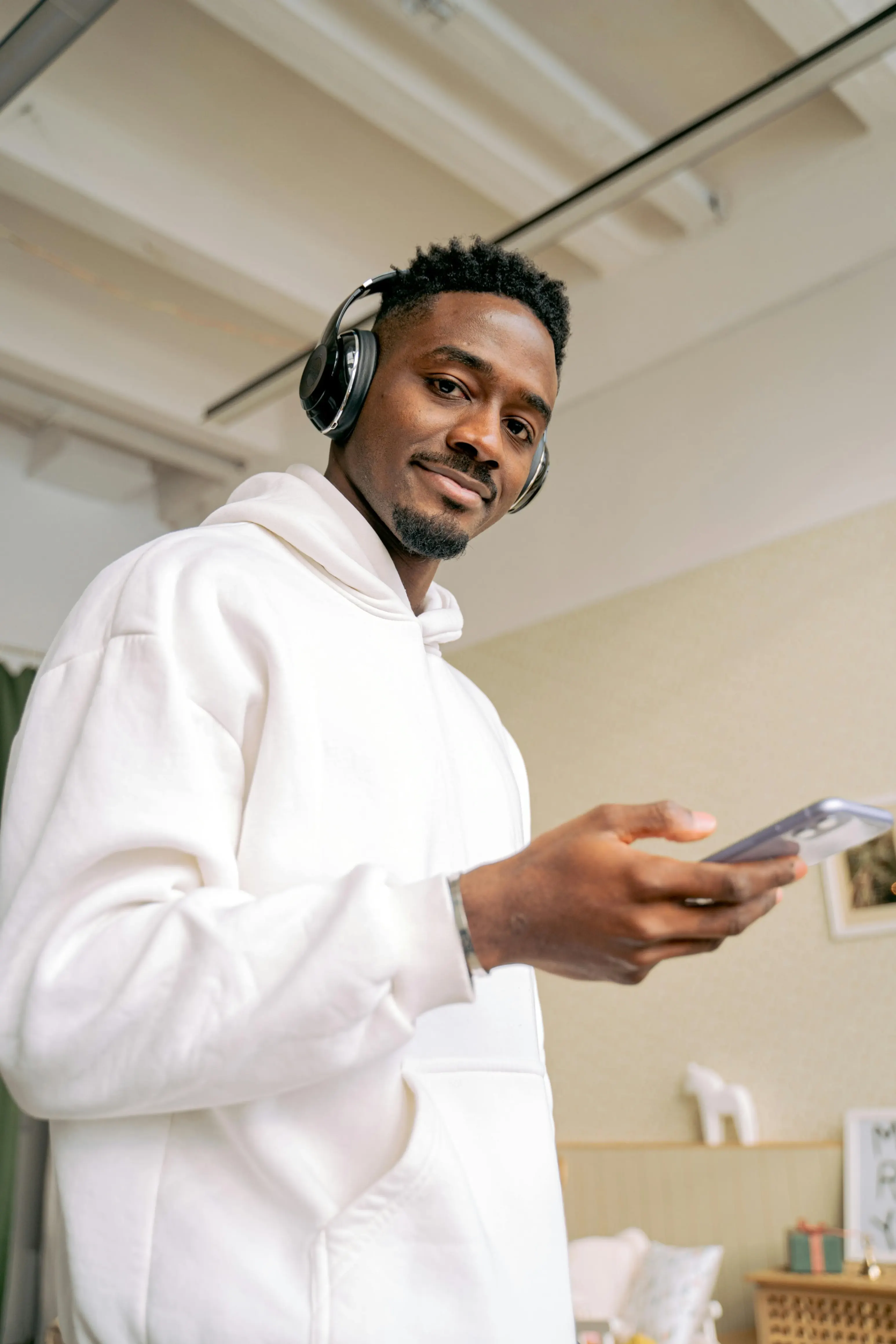 Man in white hoodie wearing headphones and holding a smartphone indoors, looking at the camera.