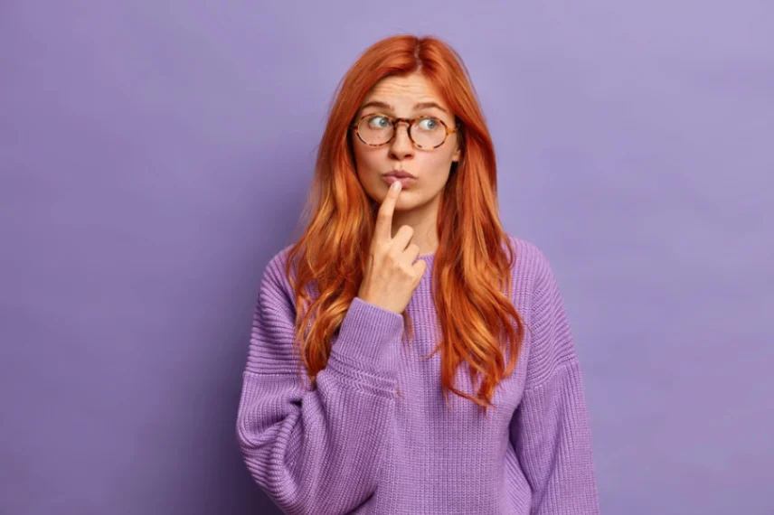 Red-haired woman wearing glasses and purple sweater, looking thoughtful with finger on lips against purple background.
