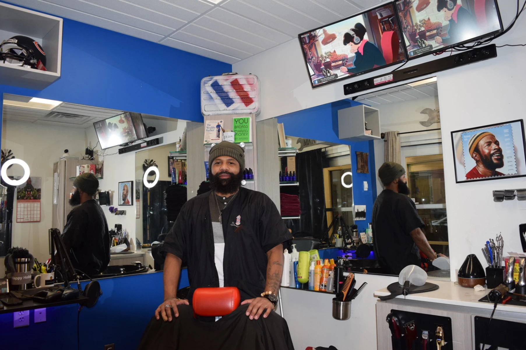 Bearded man in black barber cape sitting in a blue and white barbershop with mirrors and haircut tools.
