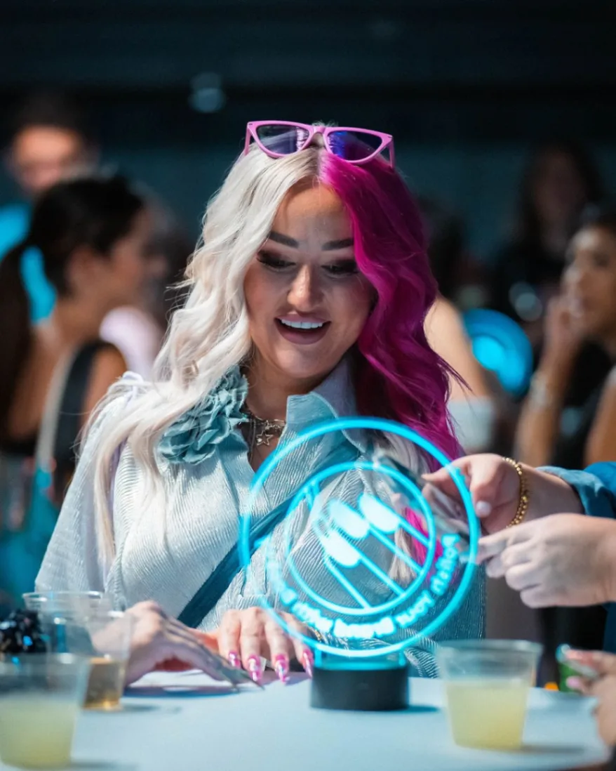 Woman with half white, half pink hair smiling at a blue glowing circular display on a table.