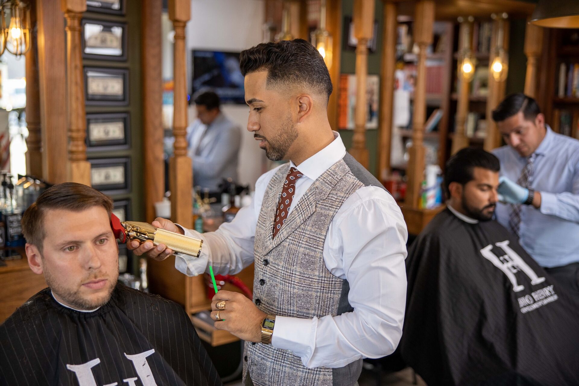 Barbers trimming hair of two seated men in a vintage-style barbershop with wood and mirrors.
