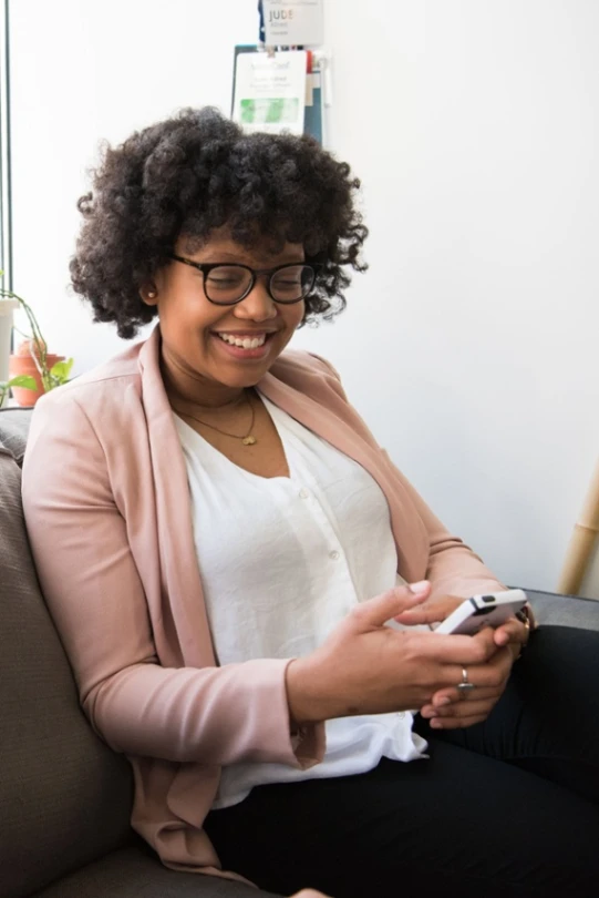 Smiling woman with glasses sitting on sofa and using phone, wearing pink blazer and white blouse.