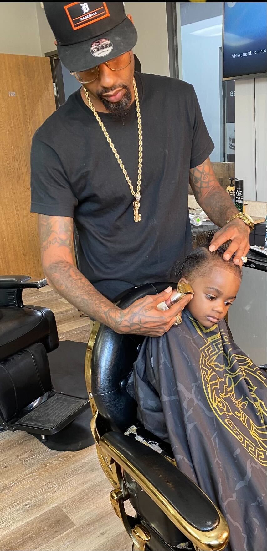 Barber with tattoos and gold chain cutting a young boy's hair in a black and gold barber chair.