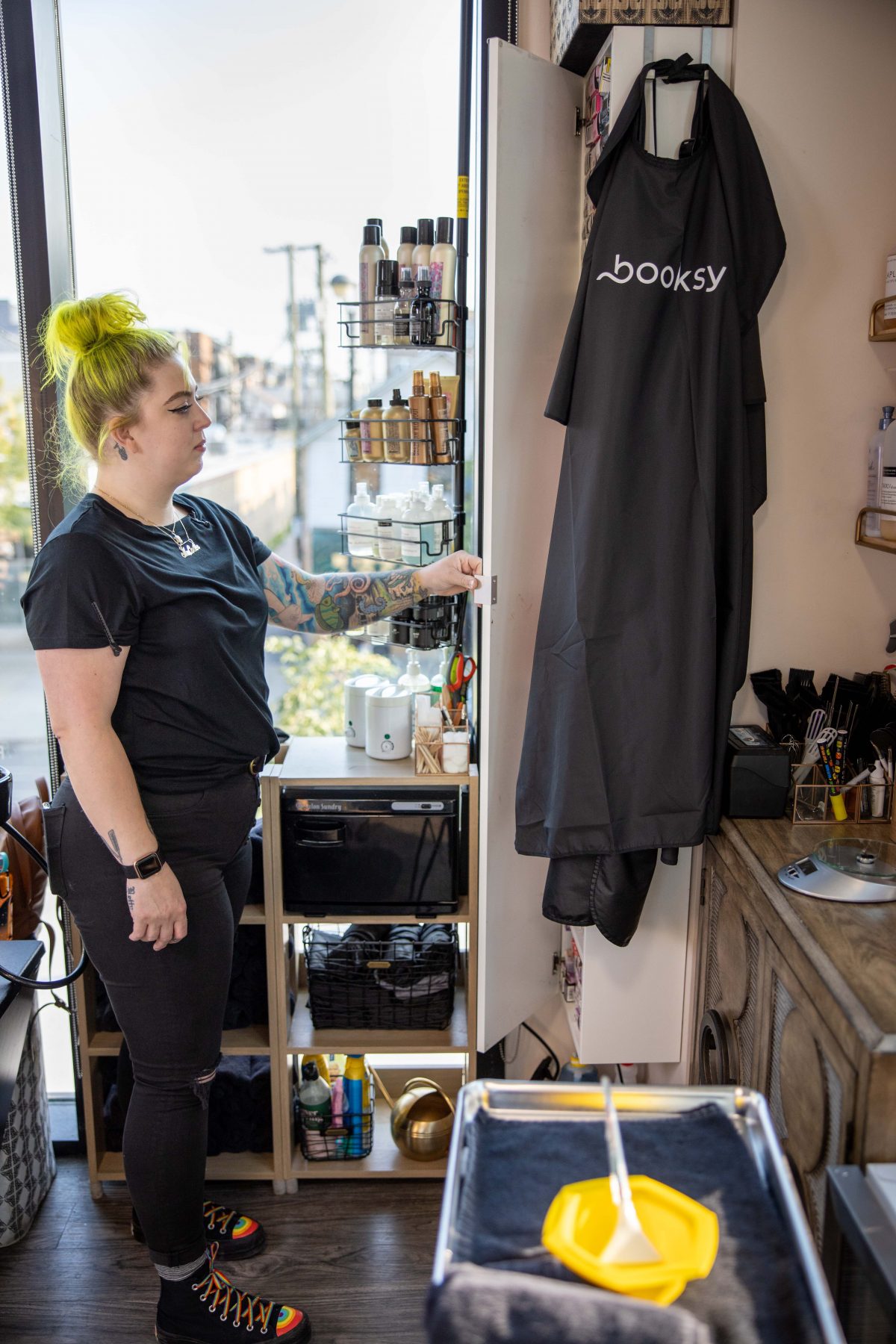Woman with yellow hair tied up stands in a salon beside shelves of hair products and a black apron.