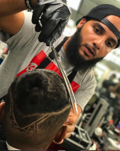 Barber with beard and cap cutting a customer's hair with scissors and gloves in a barbershop.