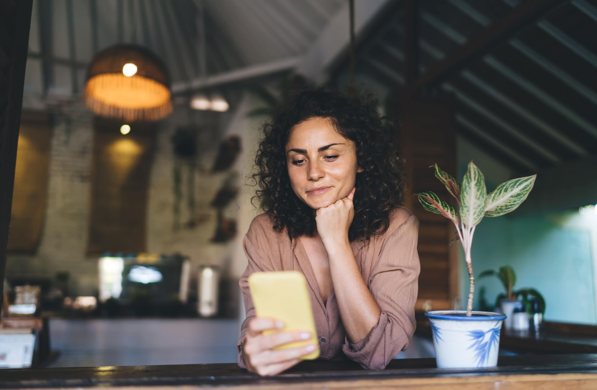 Woman with curly hair looking at a yellow phone, resting her chin on her hand at a wooden table.