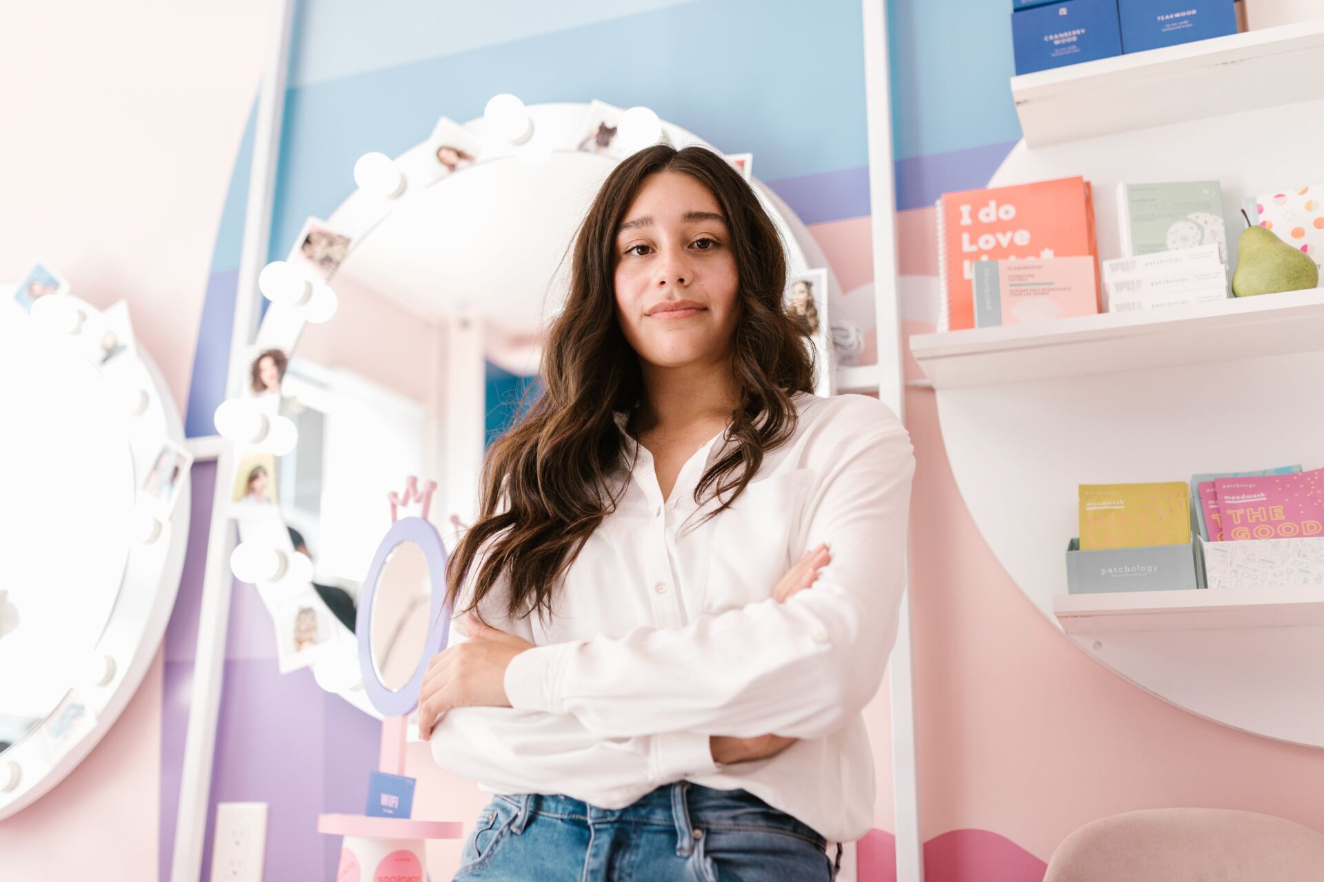 Confident woman with long dark hair in a white shirt with arms crossed standing in colorful room.