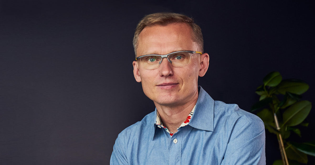 Man wearing glasses and blue shirt against a dark background with a plant to the side.