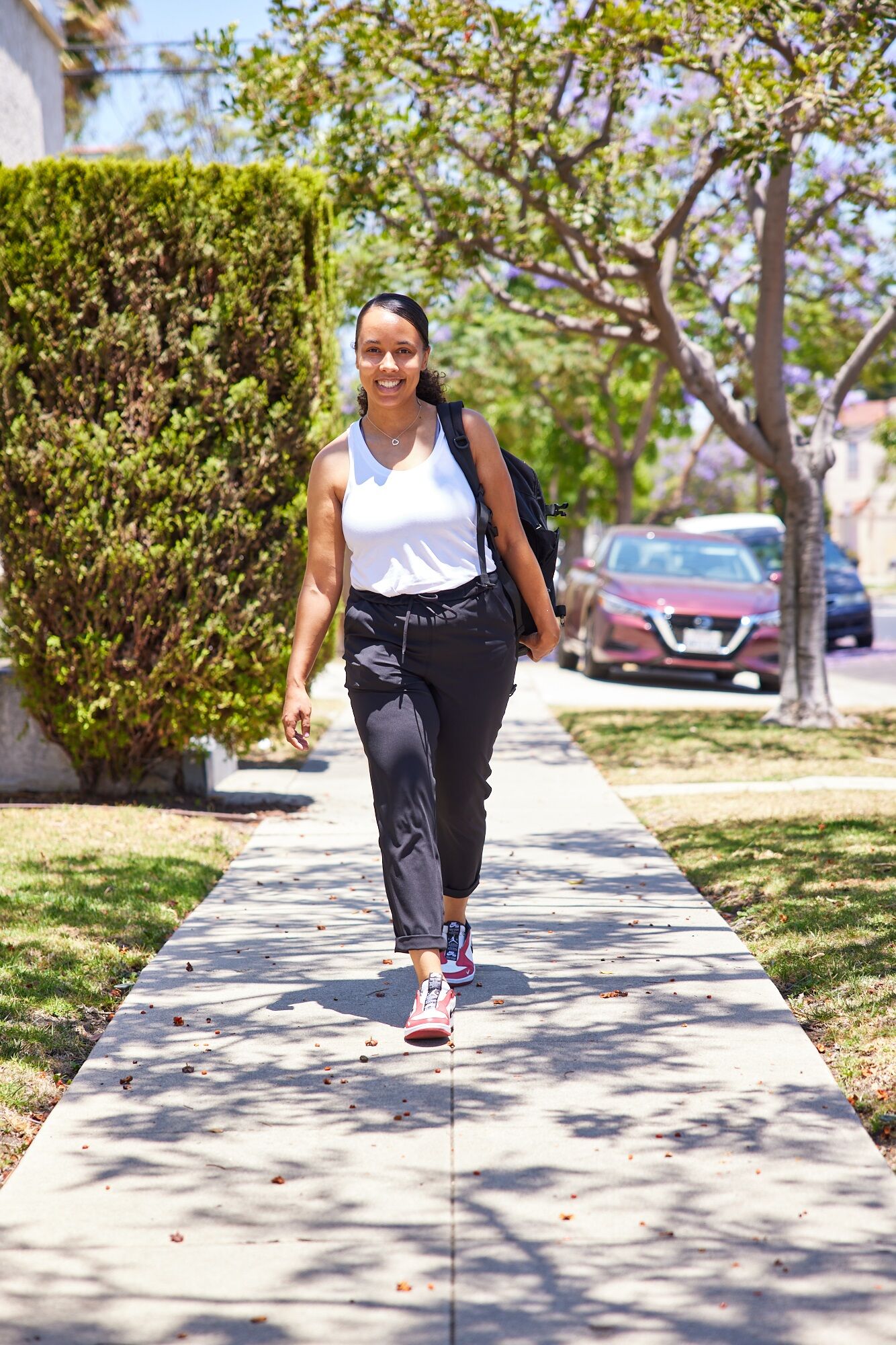 Smiling woman walking on sunny sidewalk wearing a white tank top and black pants with backpack.
