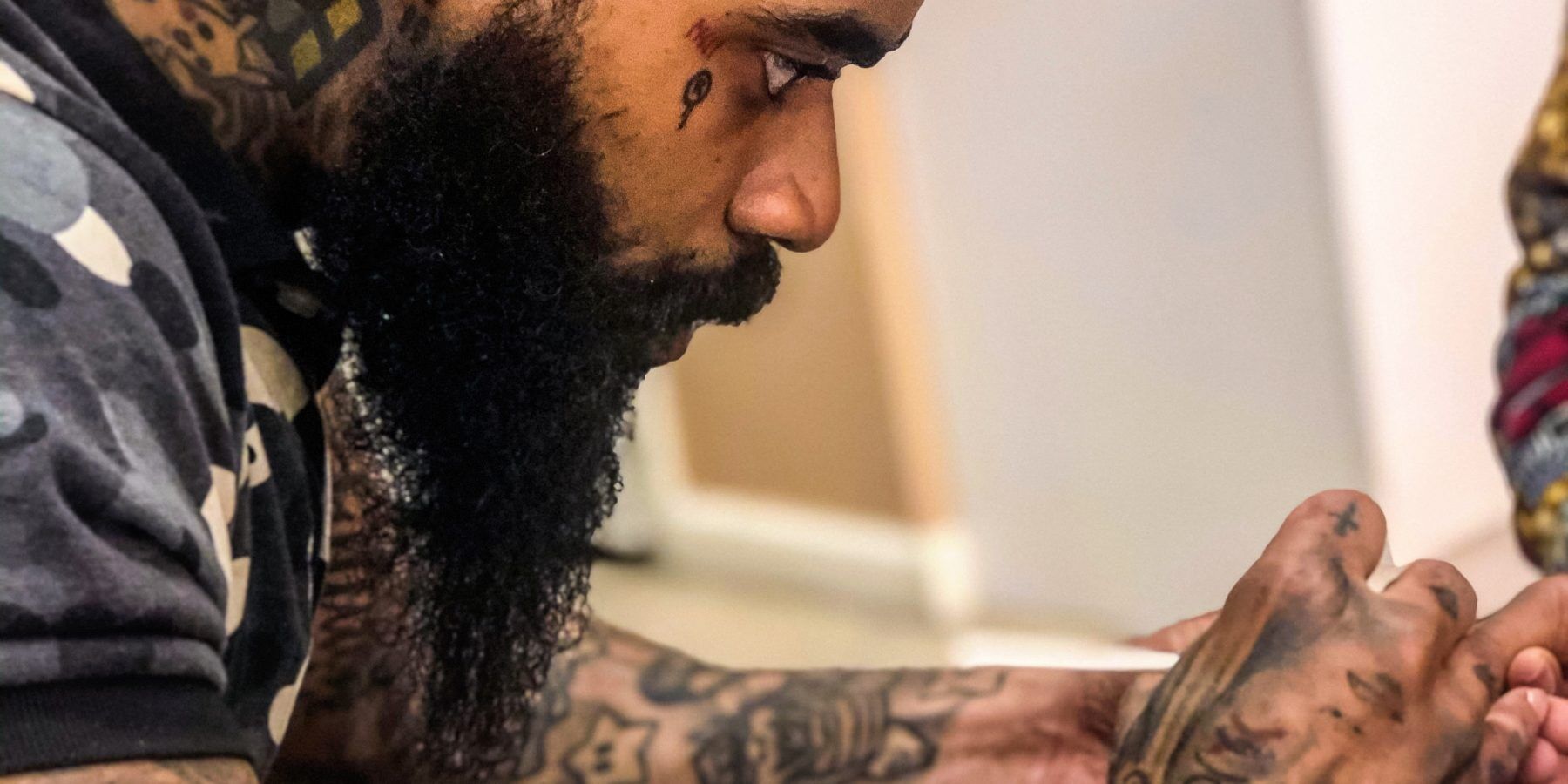Close-up of a bearded man with face and arm tattoos, focused on holding hands with another person.