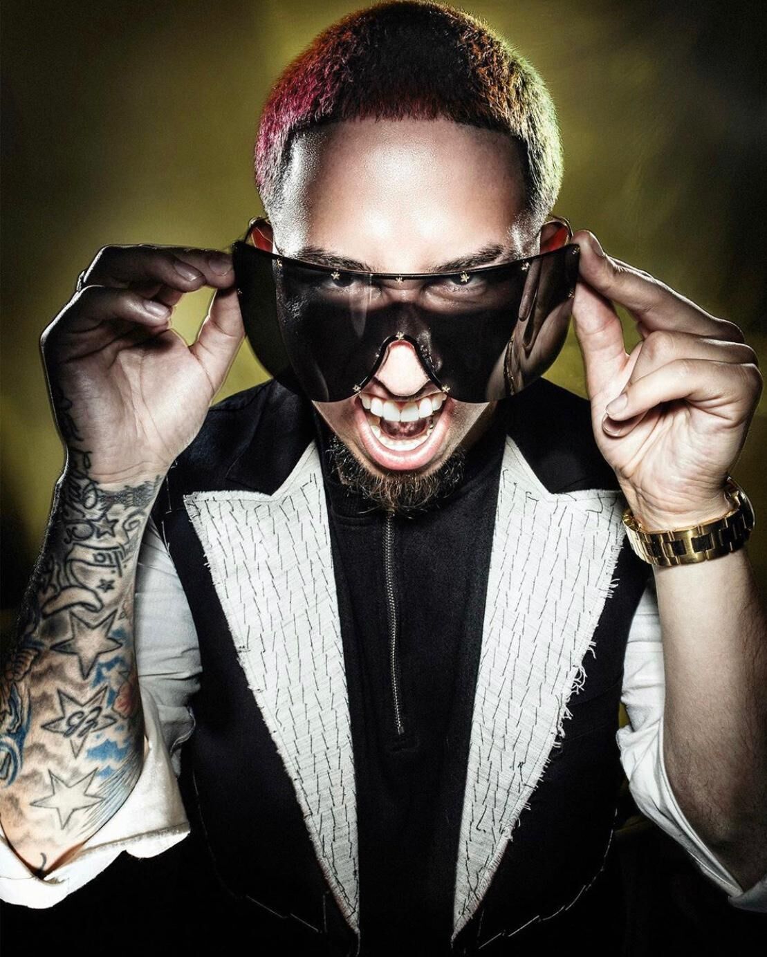Man with short hair and tattoos lifts large dark sunglasses, showing a fierce facial expression.