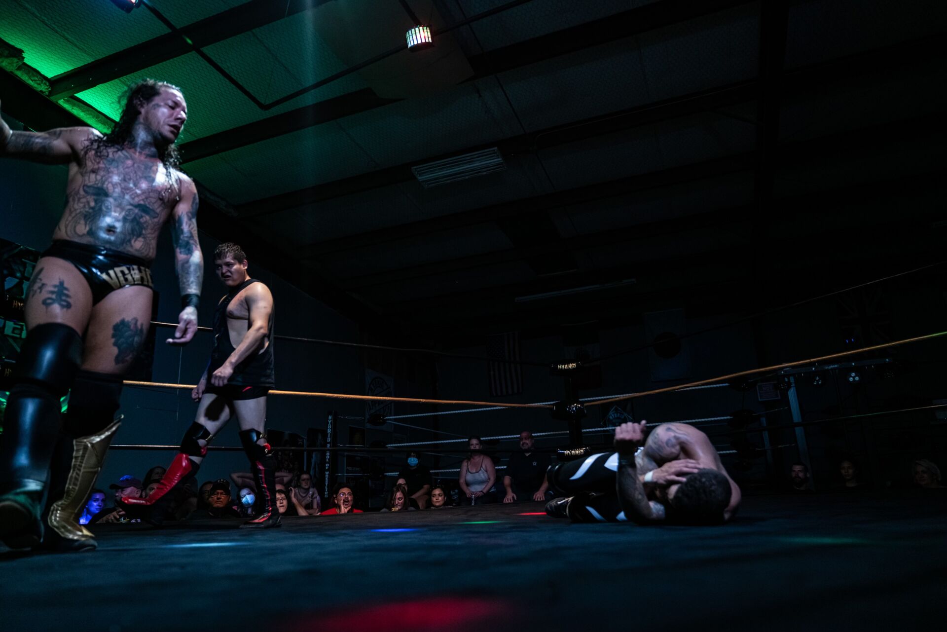 Two wrestlers standing as a third lies on the ring floor during a match with spectators watching.