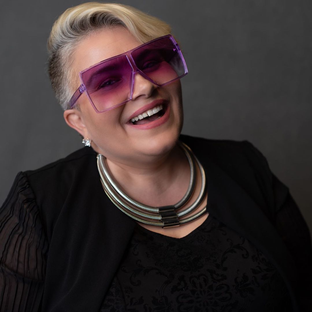 Smiling woman with short blonde hair wearing large purple sunglasses and a silver chunky necklace.