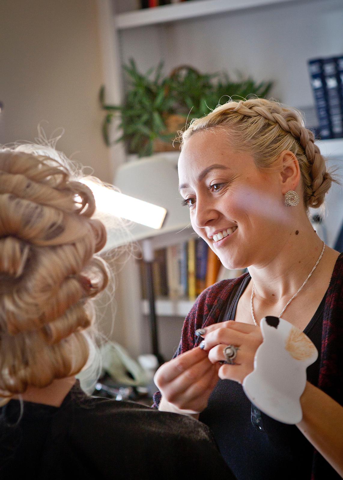 A woman with braided hair smiling while styling another person's curled hair indoors.