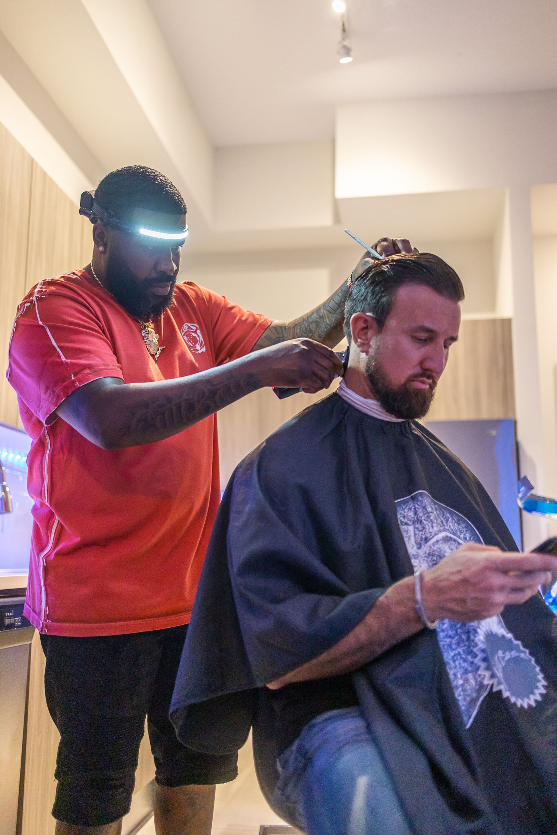 Barber wearing LED headband trims client’s hair indoors while client looks at phone.