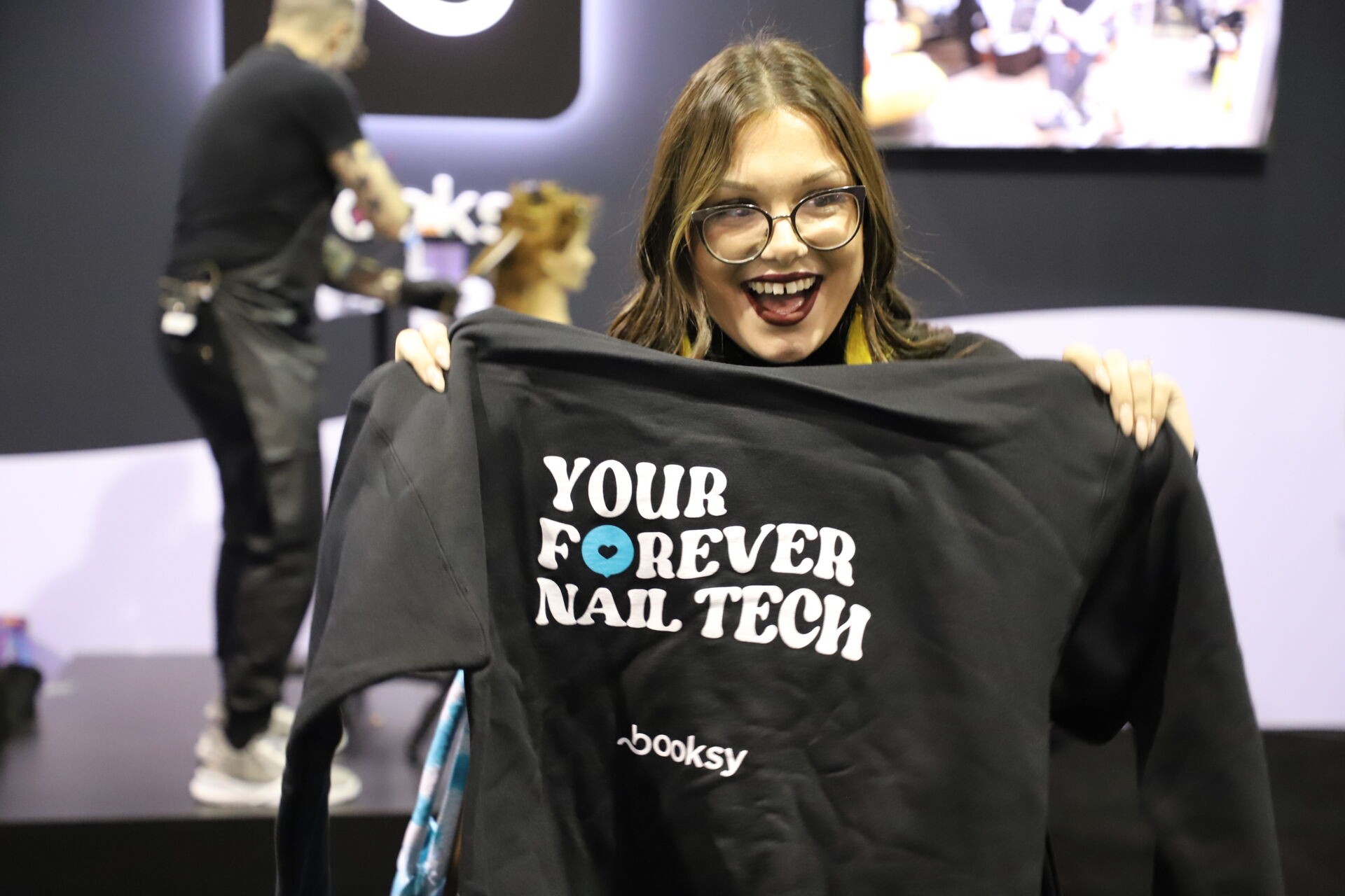 Smiling woman with glasses holds black sweatshirt that reads Your Forever Nail Tech booksy.