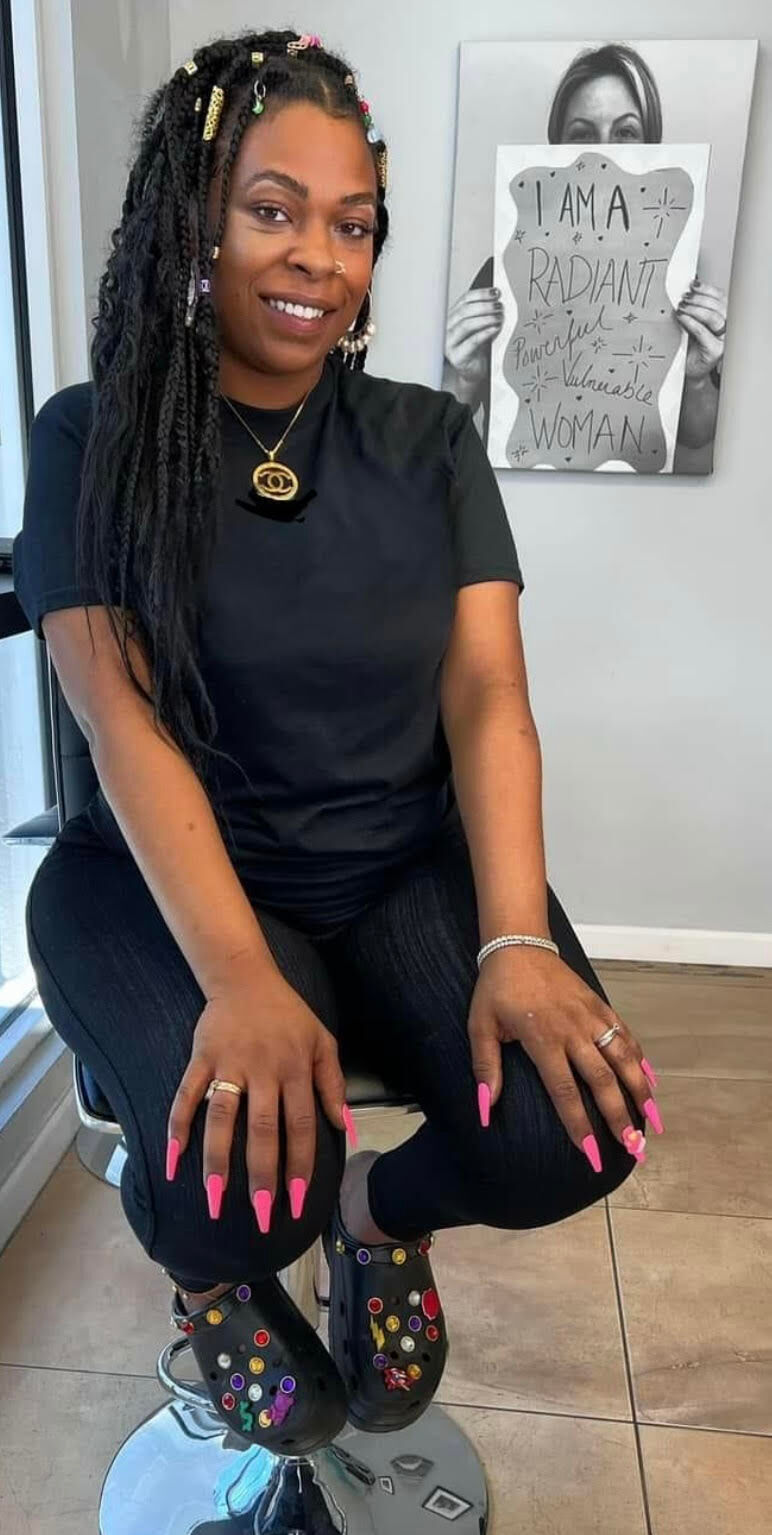 Woman with long braided hair sits on a stool, wearing black clothes and colorful decorated clogs.