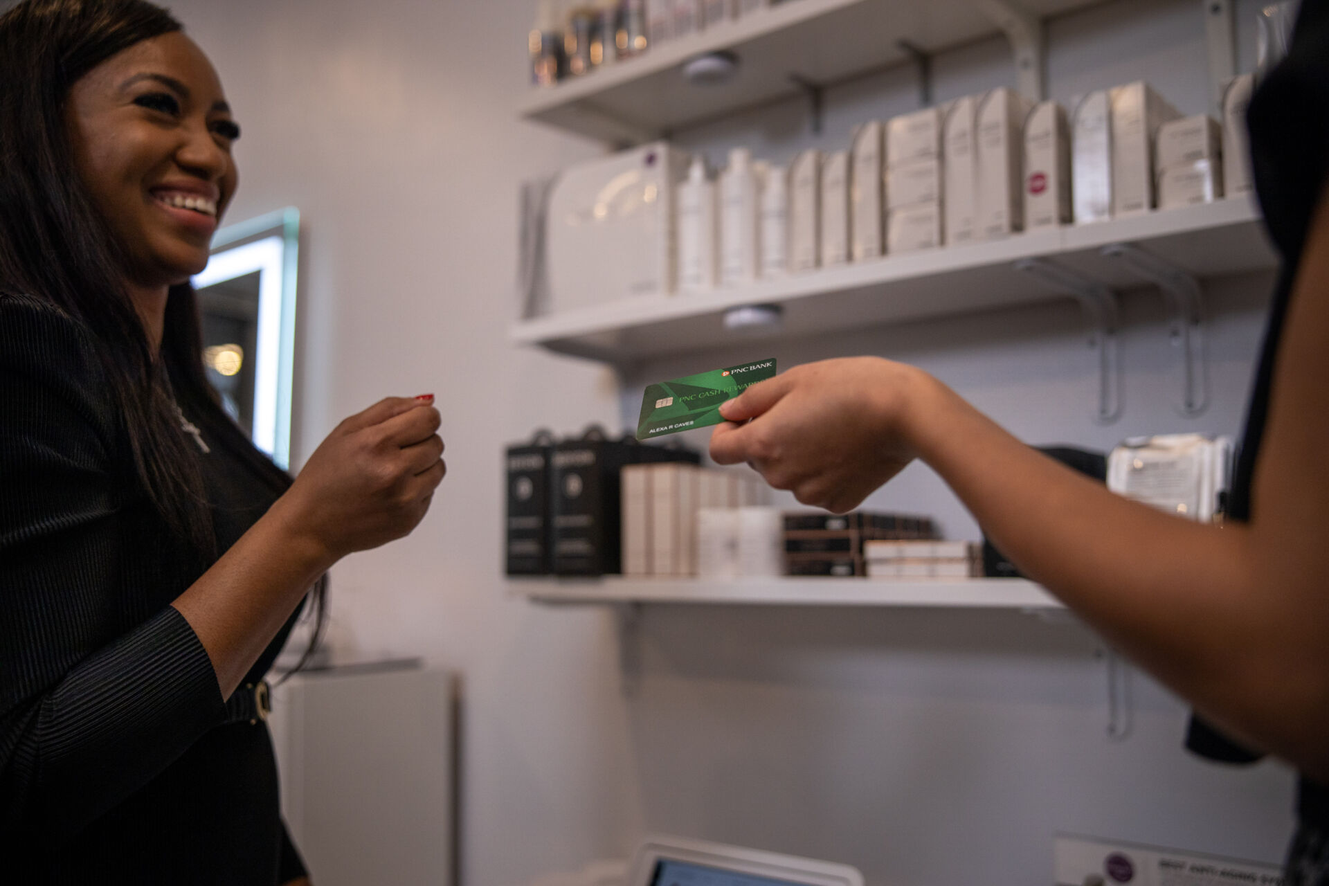 Smiling woman in black receiving a green card from another person in a store with shelves of products.